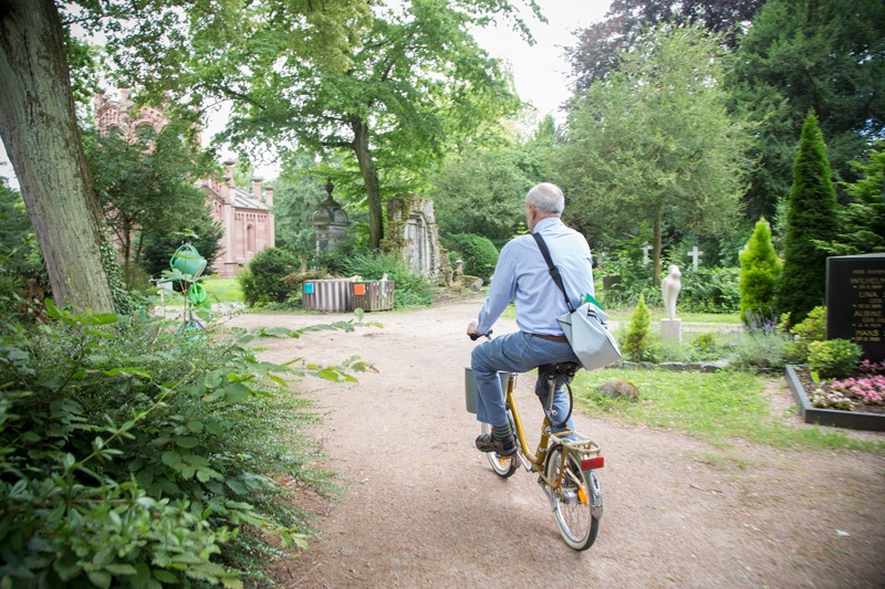 Mit seinem gelben Fahrrad fährt Georg um eine Kurve auf dem Friedhof. Seine Tasche mit dem Buch über seine Patenschaftsgräber trägt er auf dem Rücken, den Haltegriff des Eimers mit der Wurzelbürste hat er um den Fahrradlenker gehängt.