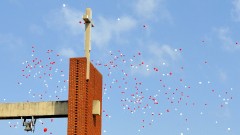 Rote und weiße Luftballons fliegen hinter einem Kirchturm mit Kreuz in den Himmel.