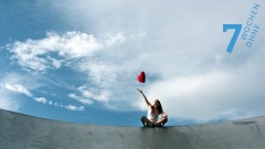 Eine junge Frau sitzt auf einer Mauer und wirft ein rotes Plüschherz in den Himmel.