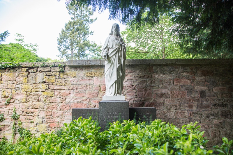 Vor einer der alten Friedhofsmauern steht eine Jesus-Figur. Grüne Pflanzen verdecken fast die Inschrift des Grabsteins.