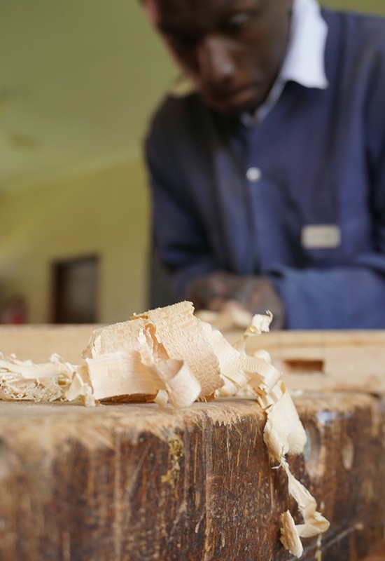 Holzspäne liegen auf einer Werkbank. Im Hintergrund arbeitet ein Schüler mit dem Hobel.