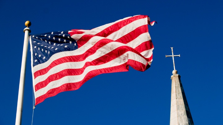 US-Flagge und Kirchturm mit Kreuz vor blauem Himmel.