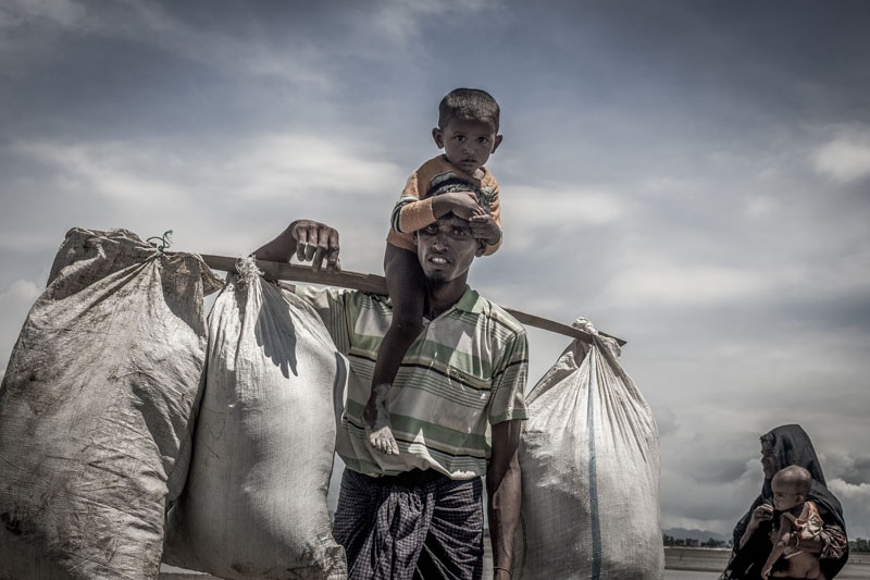Ein Rohingya-Flüchtling trägt seinen kleinen Sohnt und mehrere Säcke, die an einer Stande befestigt sind, auf den Schultern.