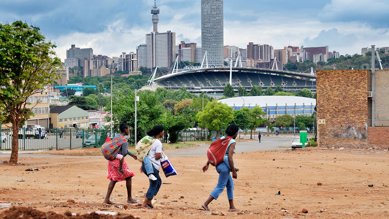 Drei junge Mütter tragen ihre Kinder auf dem Rücken. Im Hintergrund ist die Skyline von Johannesburg zu sehen. 