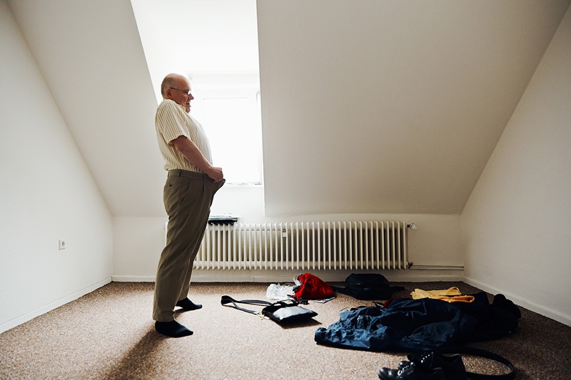 J. B. steht in einem unmöbilierten Zimmer, auf dem Boden liegen Kleidungsstücke.