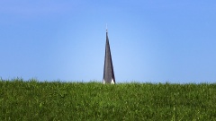 Ein Kirchturm ragt hinter einer grünen Wiese hervor.