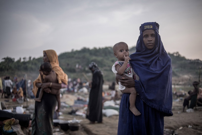 Eine Frau mit einem langen lilanen Kopftuch trägt ein Baby auf dem Arm. Sie steht vor einer Gruppe Menschen, die ihr Lager im Freien aufschlagen.