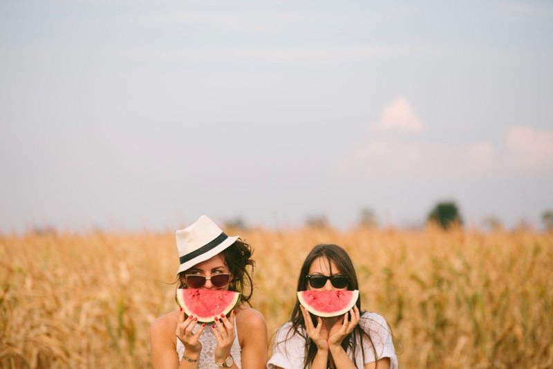 Zwei juunge Frauen sitzen auf einem Kornfeld und halten sich Melonenstücke vor den Mund.